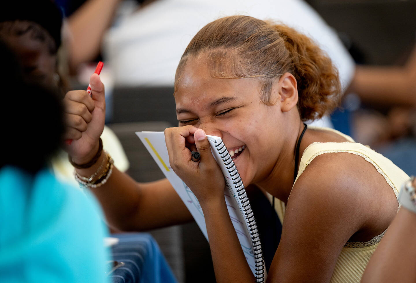 A young person laughs heartily while holding a notebook to their face, their eyes closed in joyful expression.